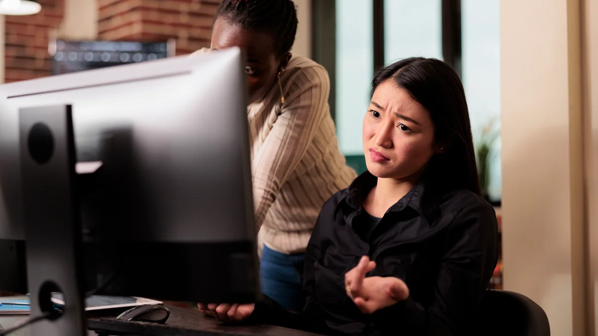 Woman confused on desktop computer alongside co-worker looking over her shoulder.